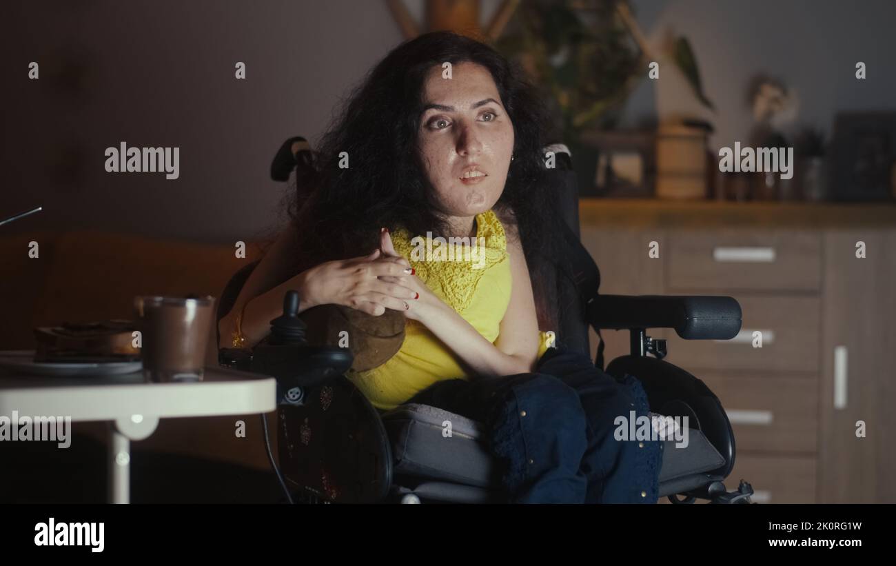 Woman with spinal muscular atrophy sitting in a wheelchair in front of TV and watching movie alone at home in the evening Stock Photo