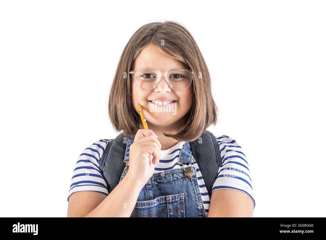 Smiling cute young schoolgirl in glasses holds pencil close to her mouth. Stock Photo