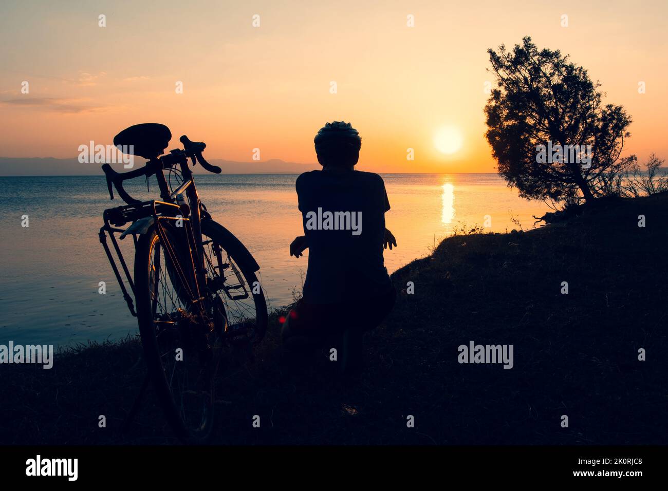 Inspirational view caucasian male cyclist sit by touring bicycle looking to scenic relaxing golden sunset background by sevan lake in Armenia. Bicycle Stock Photo