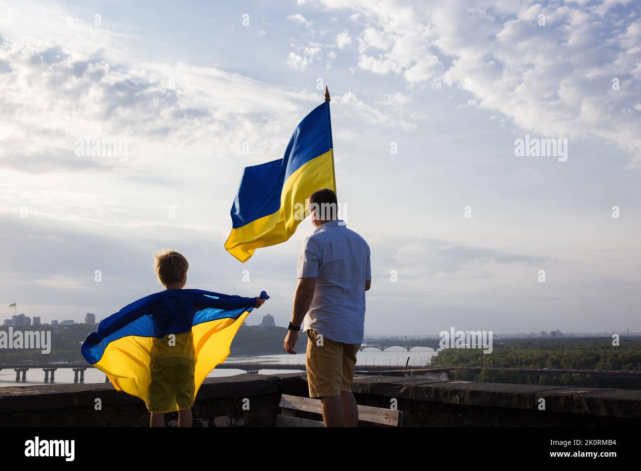 man and a child stand with their backs on the high roof of a house in Kyiv over the Dnieper river with two flags of Ukraine against the sky. Patriotic Stock Photo