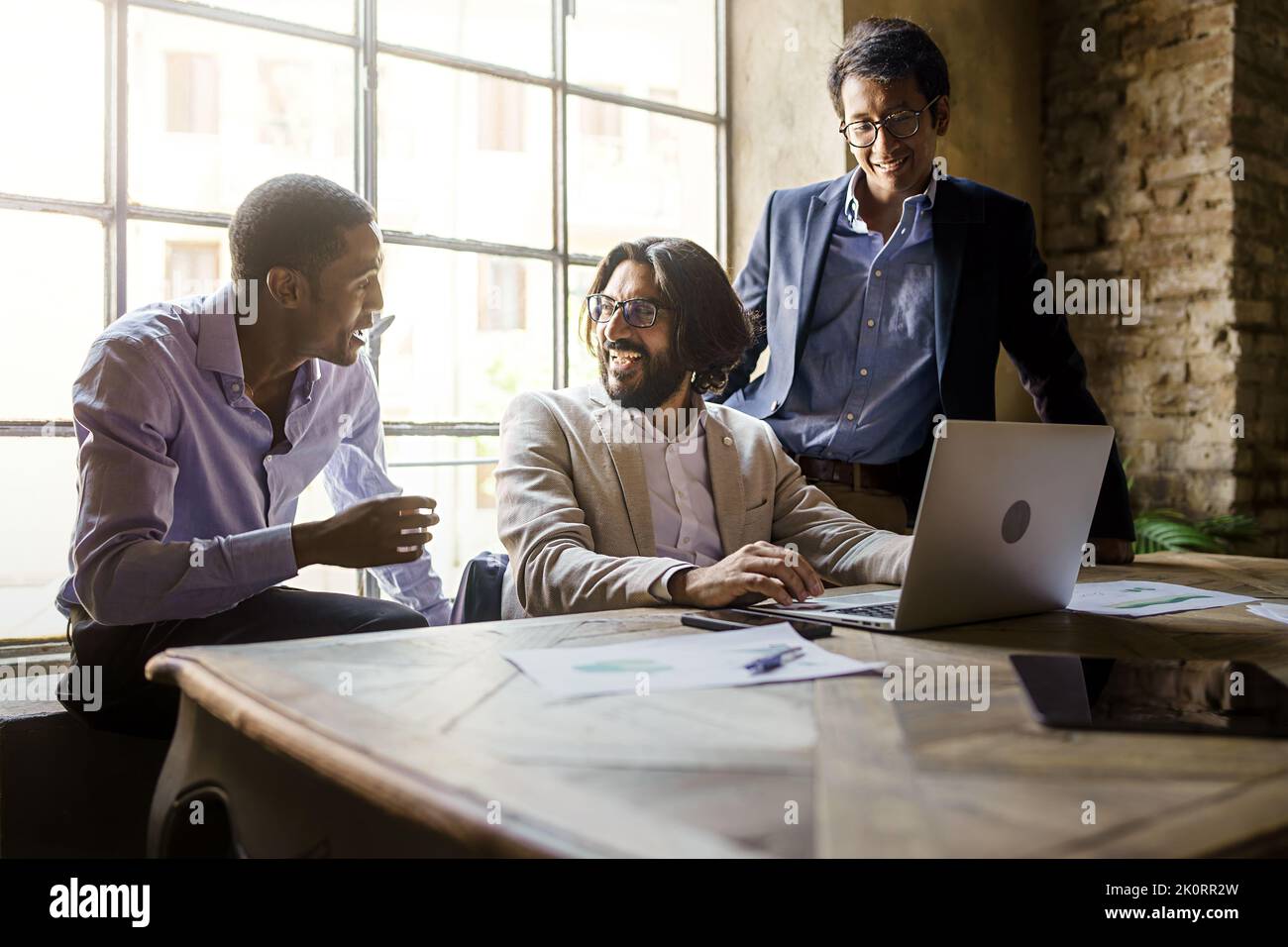 Three multiethnic young adult entrepreneurs colleagues smiling and talking about their business standing in front of a laptop computer in the office - Stock Photo