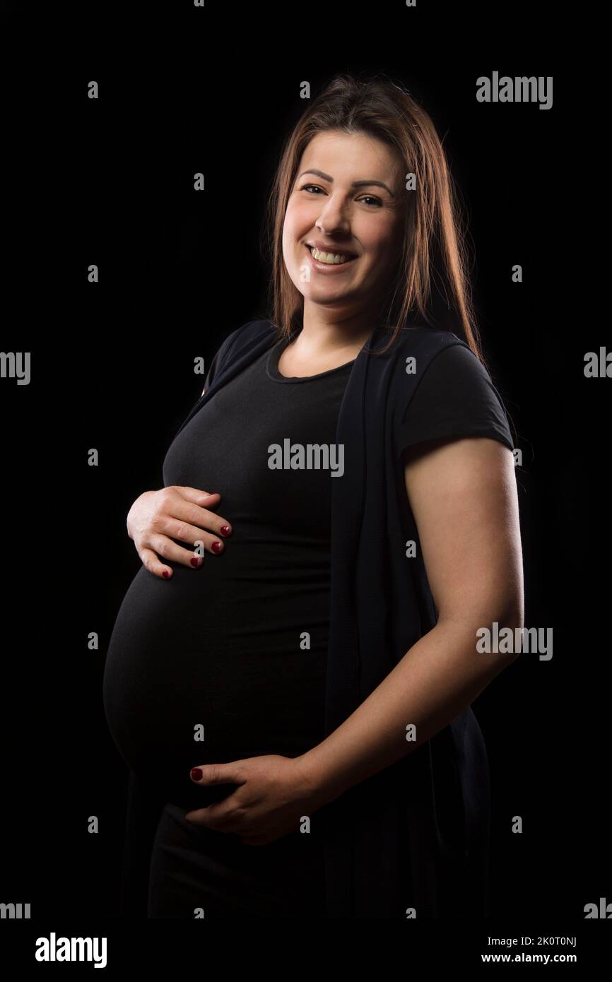 Young woman at late stages of pregnancy smiles and poses with her baby bump, studio