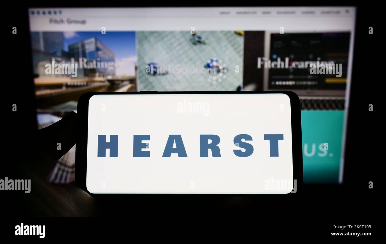 Person holding smartphone with logo of US media company Hearst Communications Inc. on screen in front of website. Focus on phone display. Stock Photo