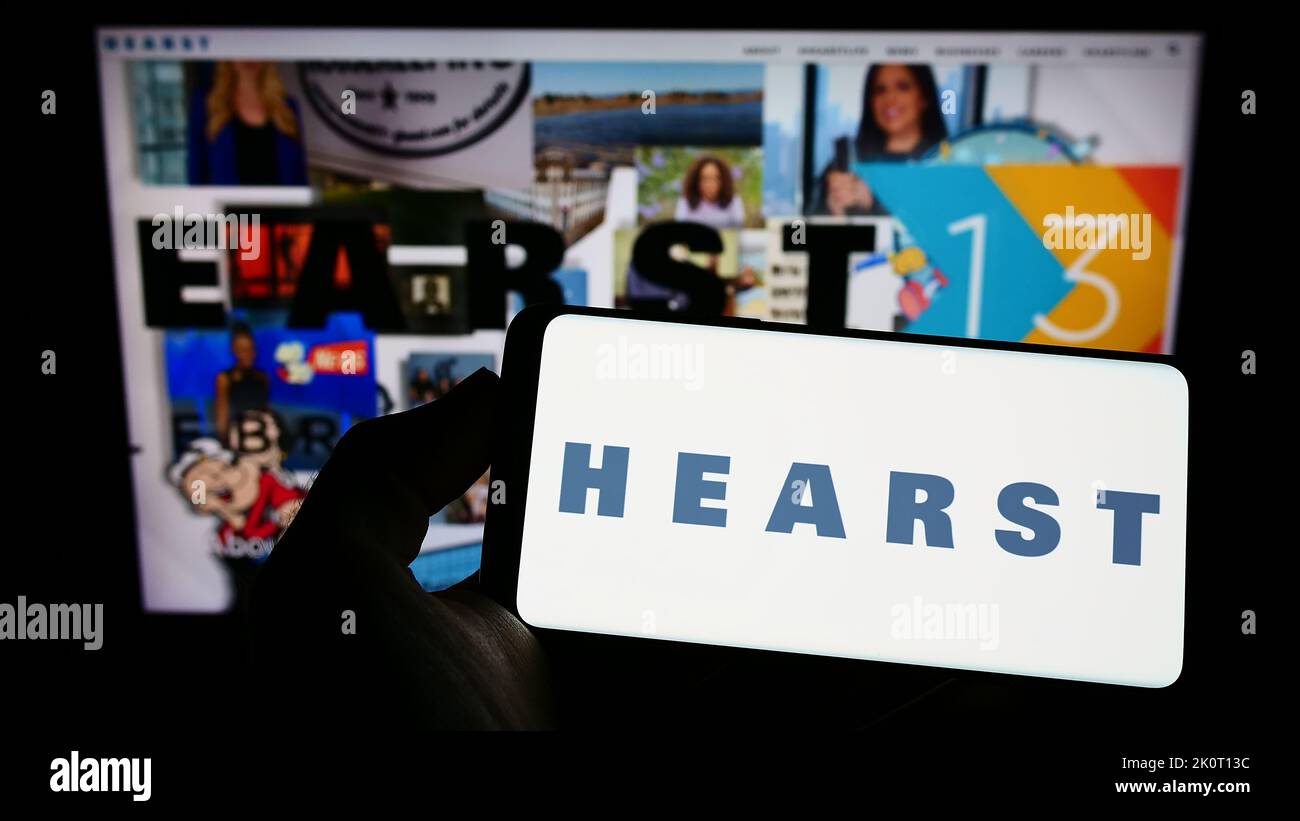 Person holding cellphone with logo of US media company Hearst Communications Inc. on screen in front of business webpage. Focus on phone display. Stock Photo