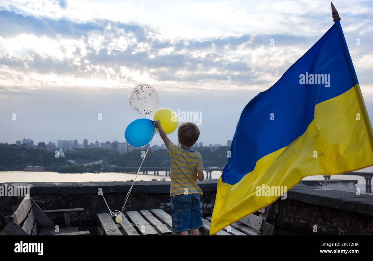boy holds yellow and blue balloons next to Ukrainian flag fluttering. stand on high roof against of city Kyiv and Dnieper River. Support Ukraine. drea Stock Photo