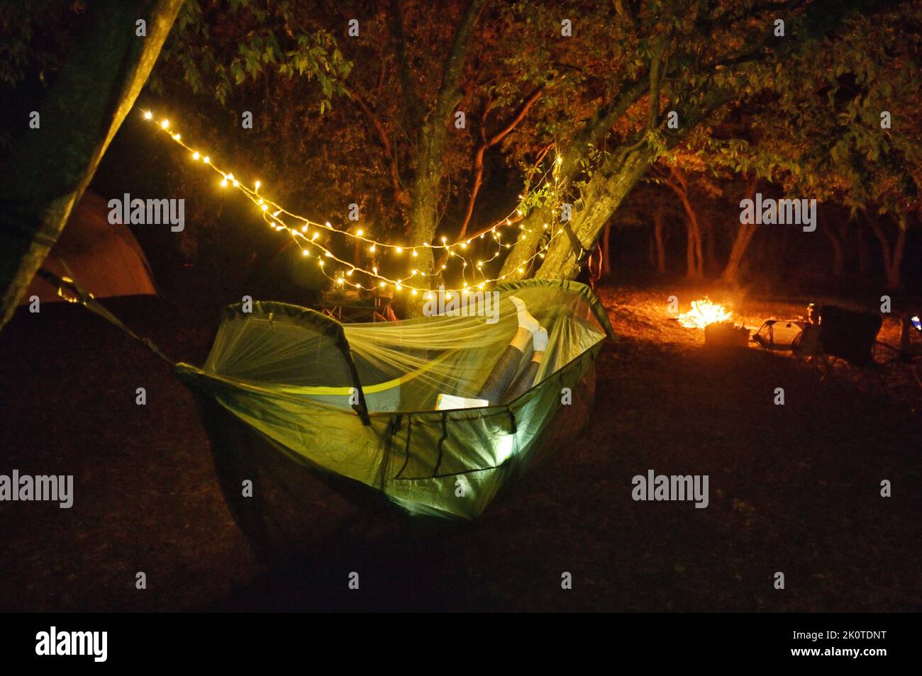 Camping in Night Forest With Sparklers. Woman Lying In Hammock and Campfire Stock Photo