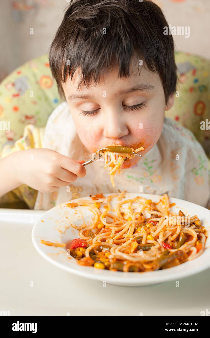 Child eating spaghetti with vegetables. Kid having fun eating. Brown