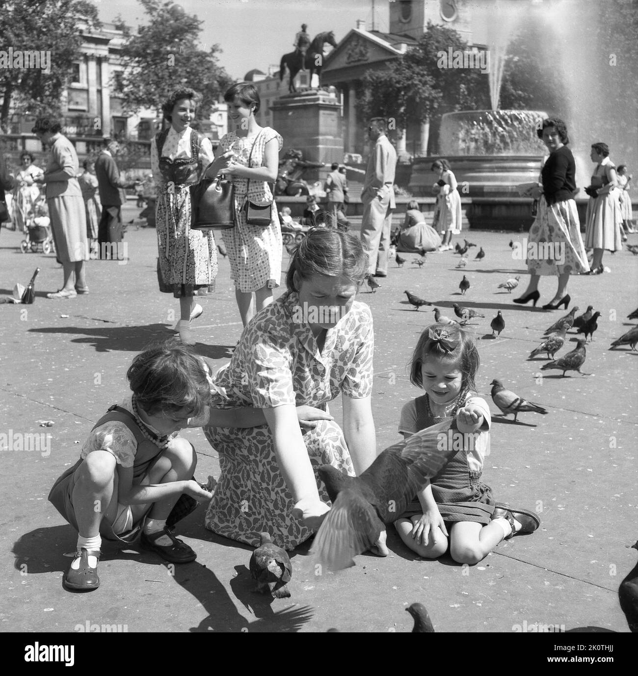 1950s, historical, with her two young daugthers beside her, a lady with her two hands outstretched feeding the pigeons at Trafalgar Square, London, England, UK. A traditional activity for over a hundred years, flocks of pigeons would descend upon the square to receive food from those visiting the famous Central London landmark. Stock Photo