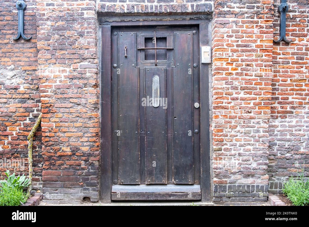 Brickwall house facade with vintage wooden door background. Doorway with irons on the door window and door phone, parterre with green plants. Netherla Stock Photo