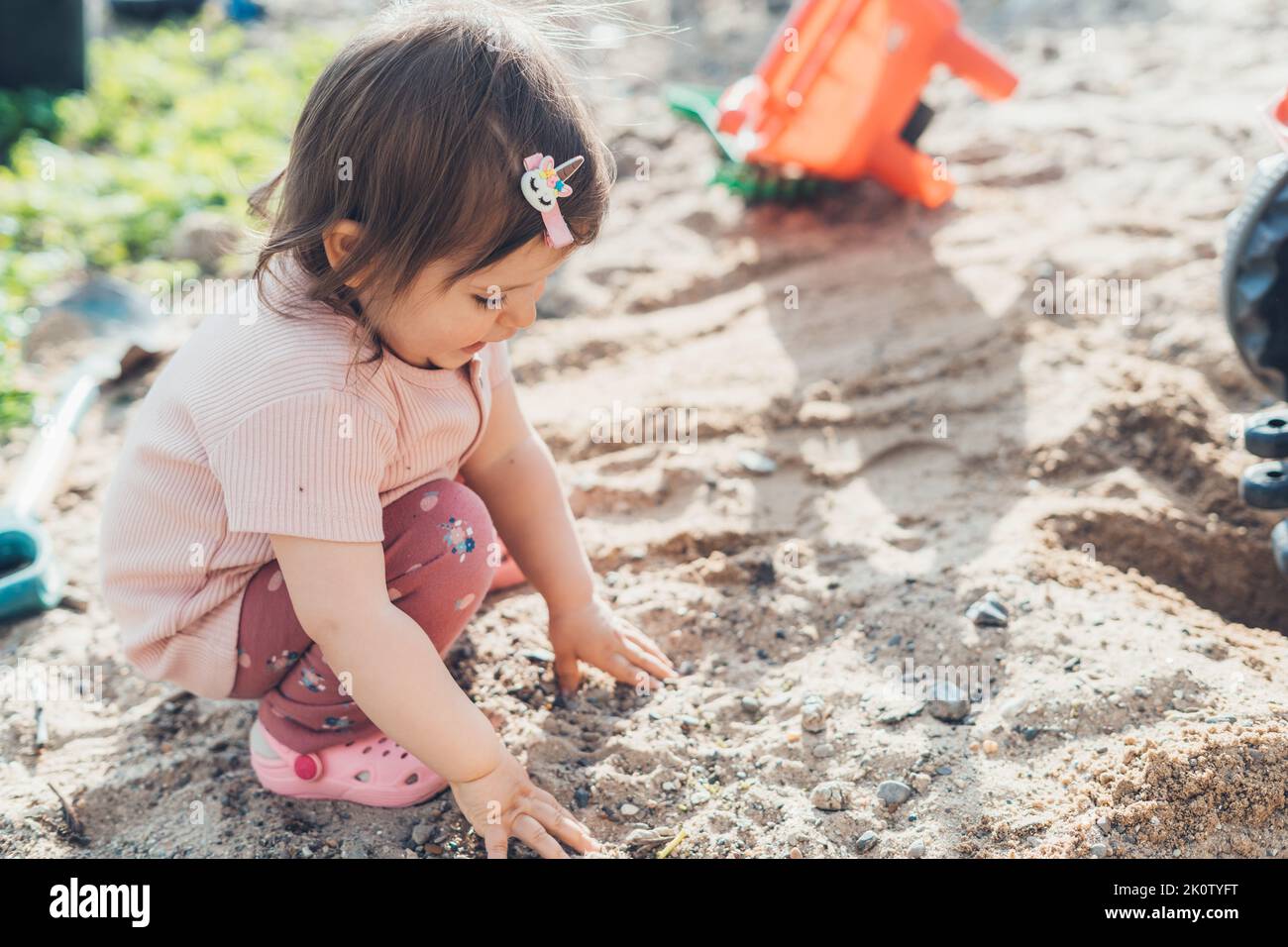 Baby girl collecting stones from the sand while playing in the yard of the house. Summer nature. Beautiful young girl. Happy family, childhood. Stock Photo