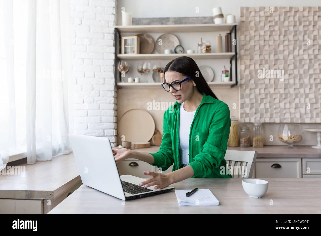 Deadline. A beautiful young business woman in glasses sits at the table in the kitchen, works with a broken laptop, a computer malfunction, waves her hands unhappily, points to the monitor. Stock Photo