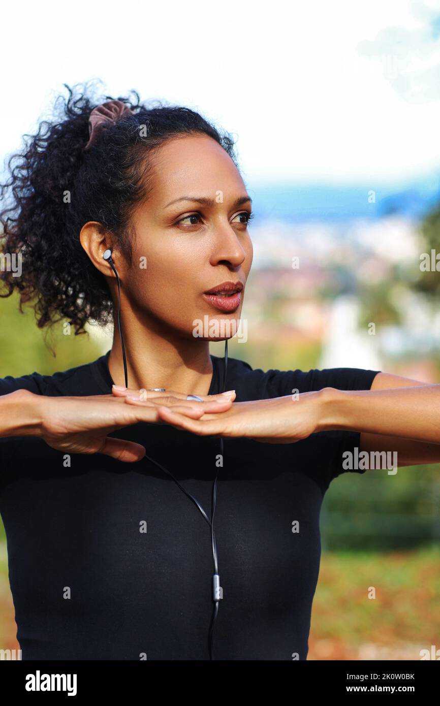 close up woman outdoors with earphones and curly hair hands on chest sports outfit Stock Photo