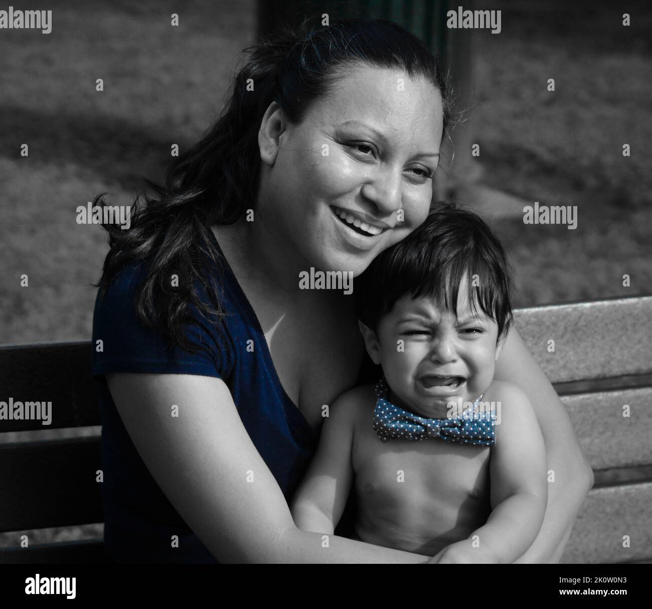 A smiling latina mother holds her crying baby on a park bench Stock Photo