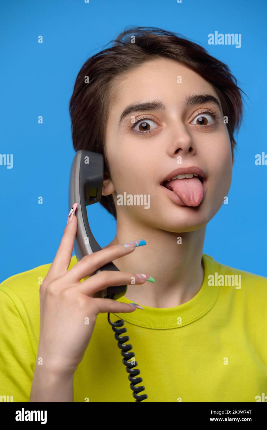 Beautiful young skinny brunette grimaces with tongue out while talkng on landline phone Stock Photo