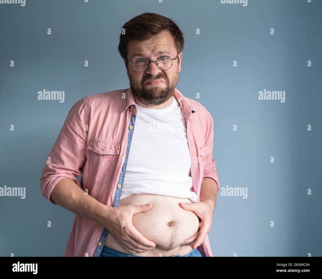Unhappy, dissatisfied overweight man, lifted t-shirt and holds excess belly fat with his hands. Healthy lifestyle concept. Stock Photo