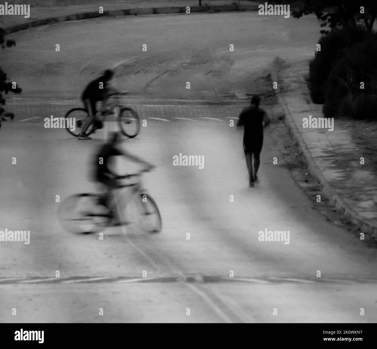 A grayscale shot of a jogger running along an empty curving road with two bicycle riders following him Stock Photo