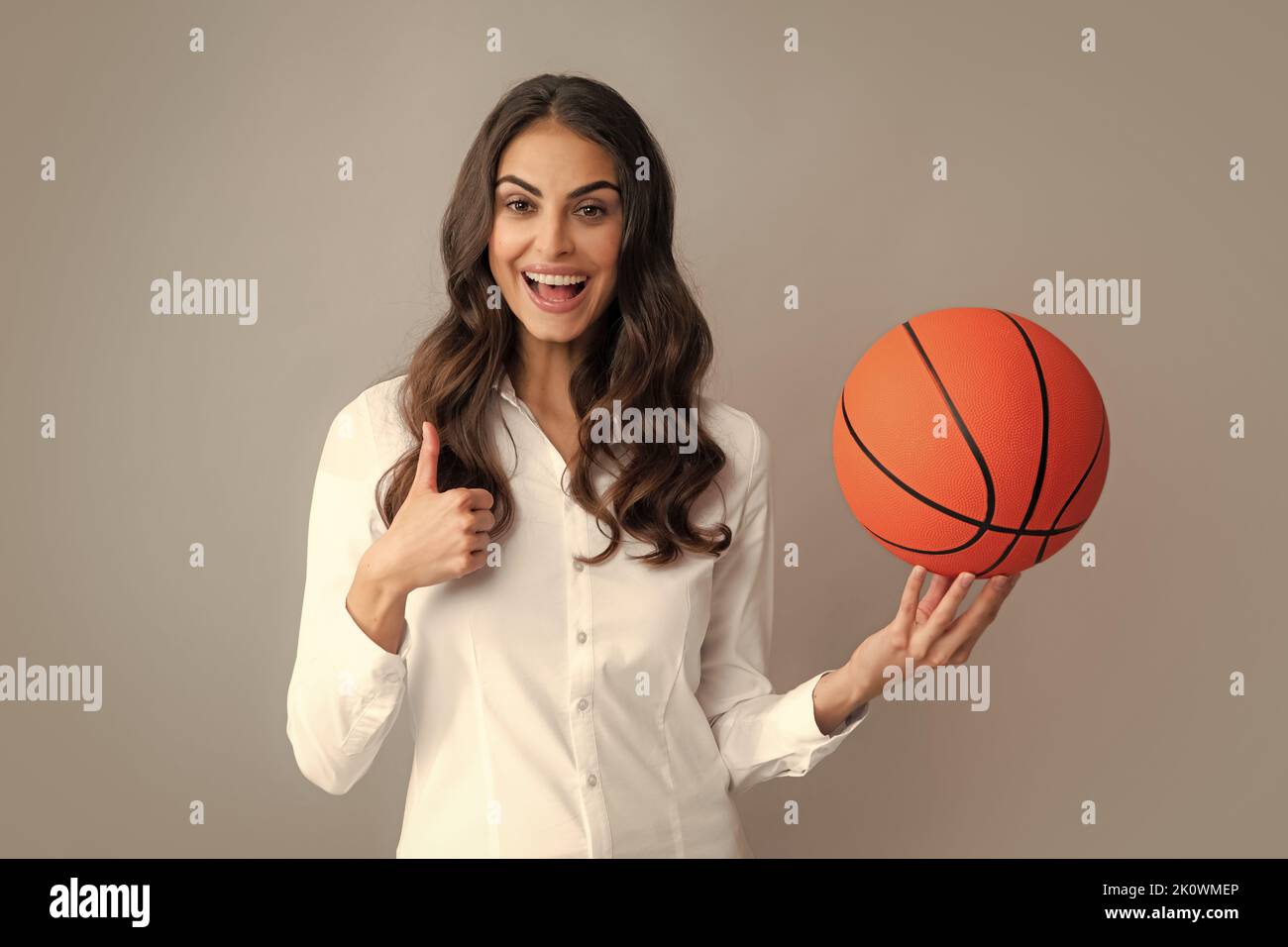 Happy woman with thumb up holding a basketball ball, isolated on gray background. Stock Photo