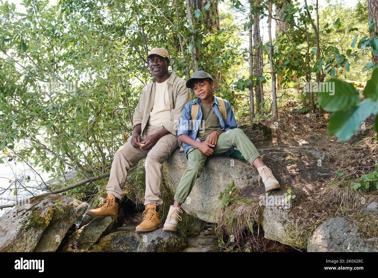 Restful mature black man and his grandson in activewear enjoying halt on huge stone covered with moss and dry grass in the forest Stock Photo