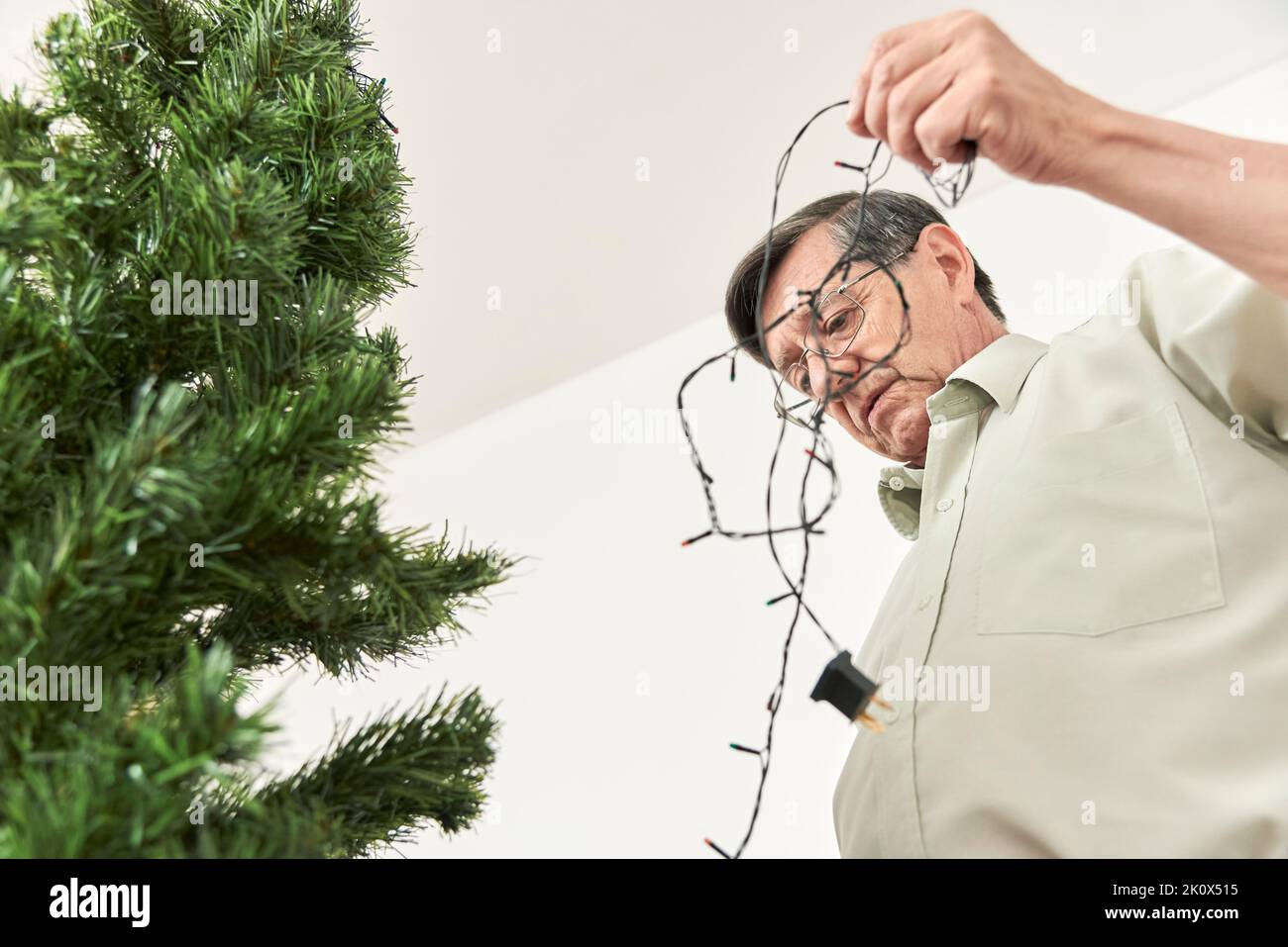 Concern about the use of electrical energy during the holidays. Hispanic senior man stretching a string of lights to put on a Christmas tree in his ho Stock Photo