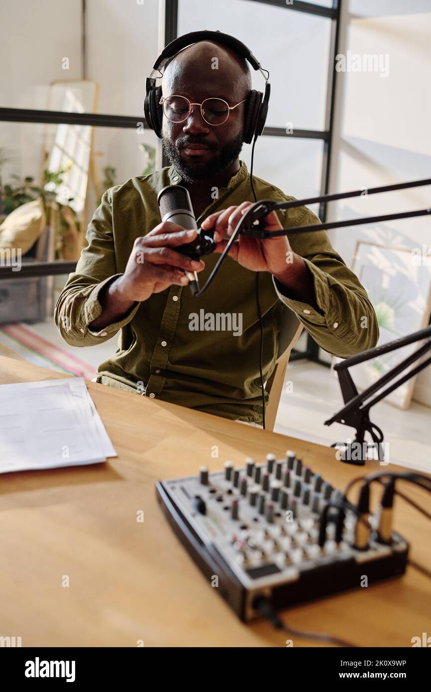 Young serious black man in headphones and eyeglasses adjusting microphone before recording new audio file for online audience Stock Photo