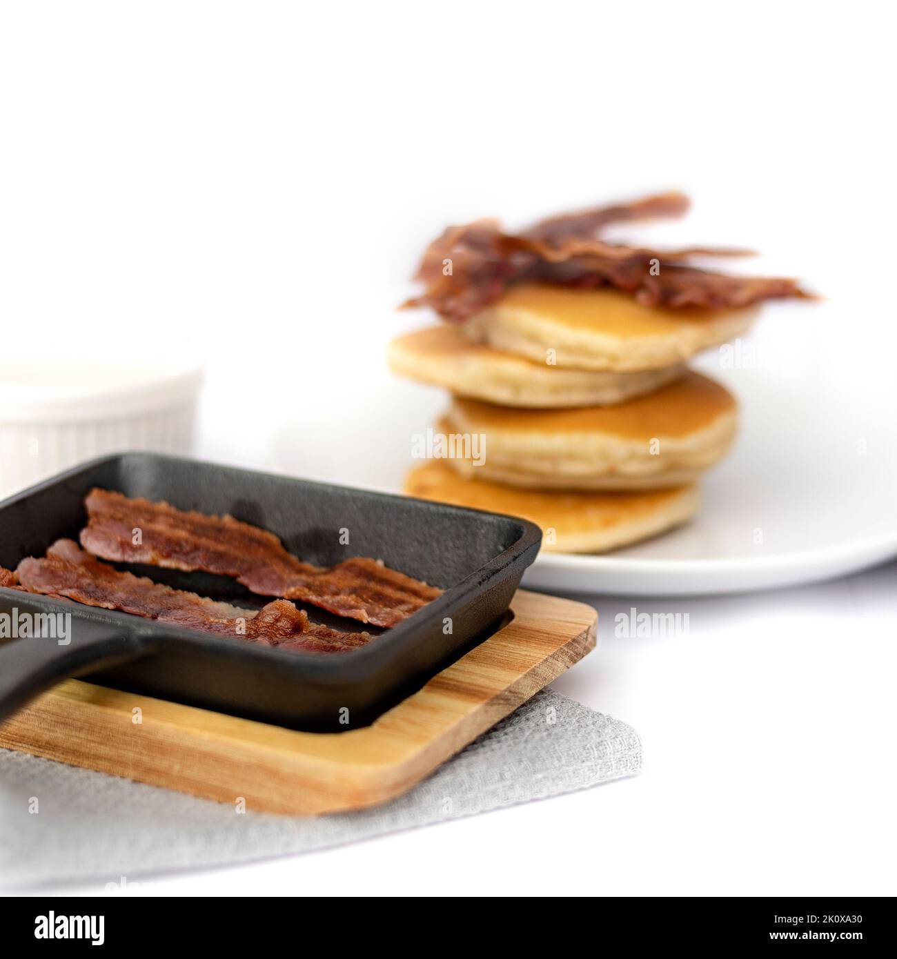Mini cast-iron skillet with crispy bacon and pancake stack in the background isolated against white background Stock Photo