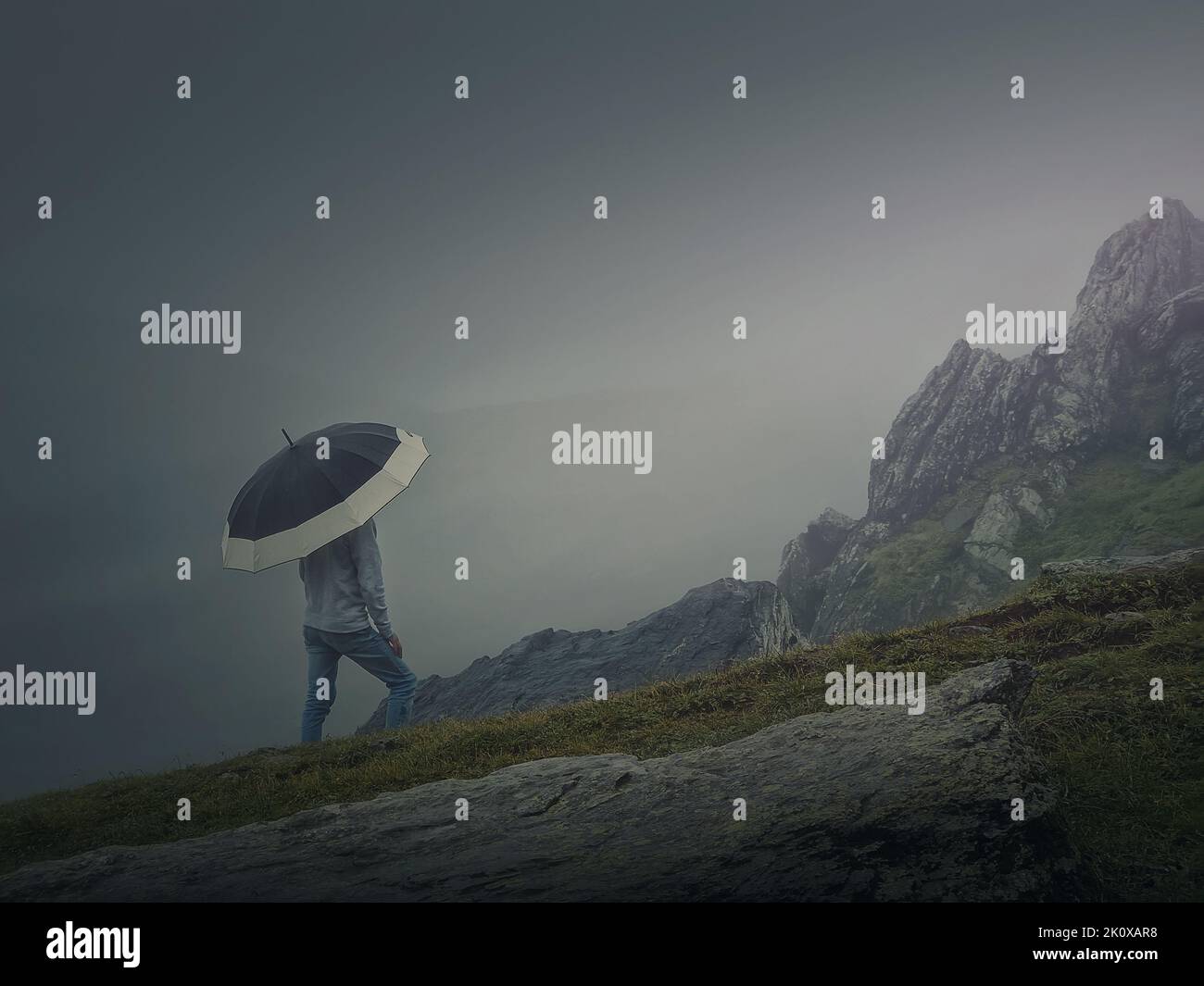 Rear view of a lonely man with umbrella stands on a rocky hill covered by haze. Moody and emotional scene with a lone stranger silhouette under rain Stock Photo