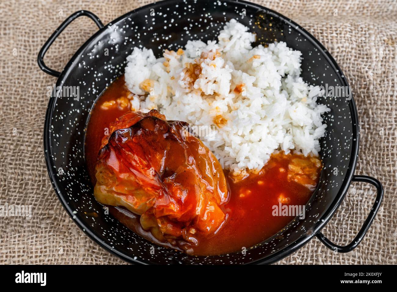 Baked stuffed paprika with minced beef in tomato sauce with stewed rice on black rustic pan on jute fabric background. Stock Photo