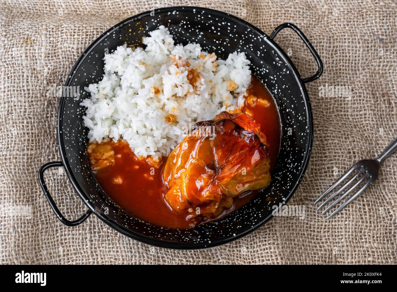 Baked stuffed paprika with minced beef in tomato sauce with boiled rice on black rustic pan on jute fabric background with fork. Stock Photo