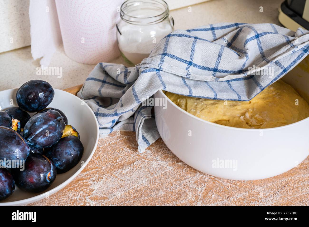 Plum on plate, yeast dough in bowl with towel, glass with sugar and paper kitchen towel on wooden board sprinkled with flour, closeup. Preparing sweet Stock Photo