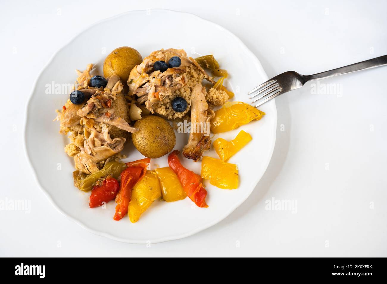 Baked piece of hen meat on couscous, potato, baked multicoloured paprika and blueberry on white plate, fork on white background, closeup. Serving size Stock Photo