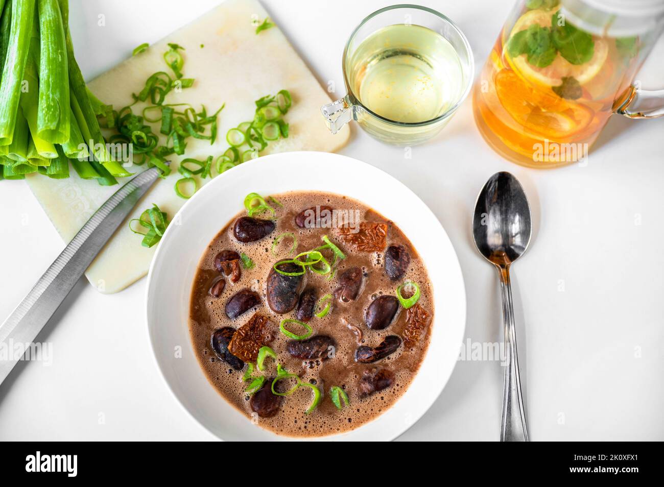 Giant bean soup with dried tomato in plate, sliced green onion shoot on kitchen board, orange fruit beverage, spoon and knife on white background. Stock Photo