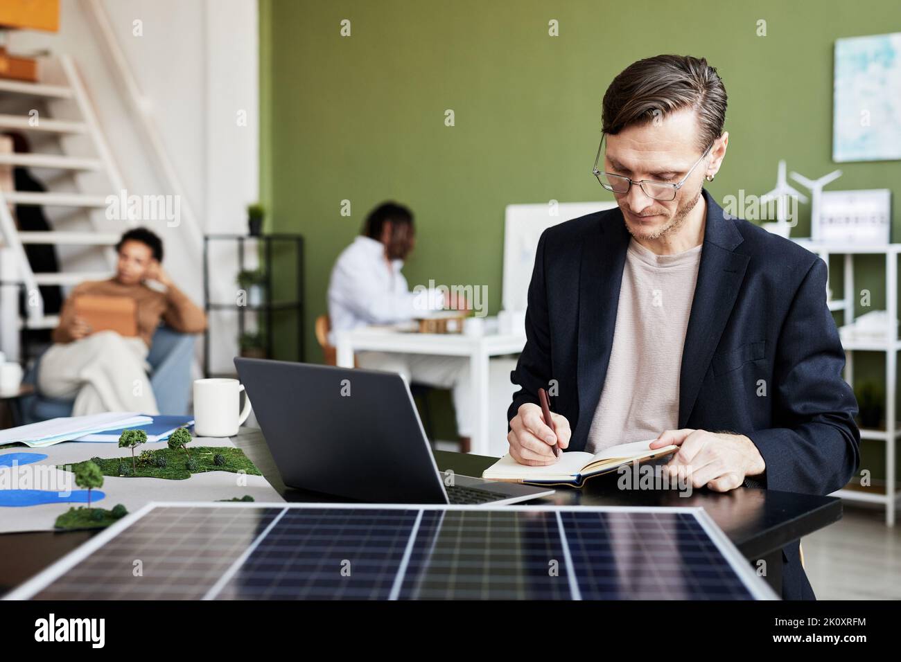 Young businessman making notes in notebook while working at table with laptop and solar panel Stock Photo