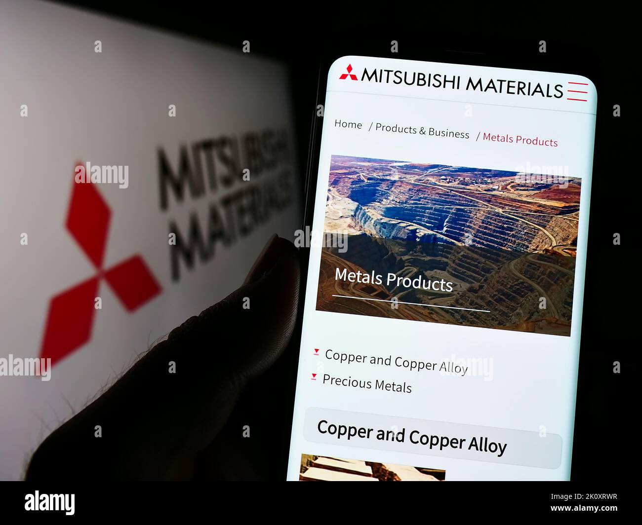Person holding smartphone with website of company Mitsubishi Materials Corporation (MMC) on screen with logo. Focus on center of phone display. Stock Photo