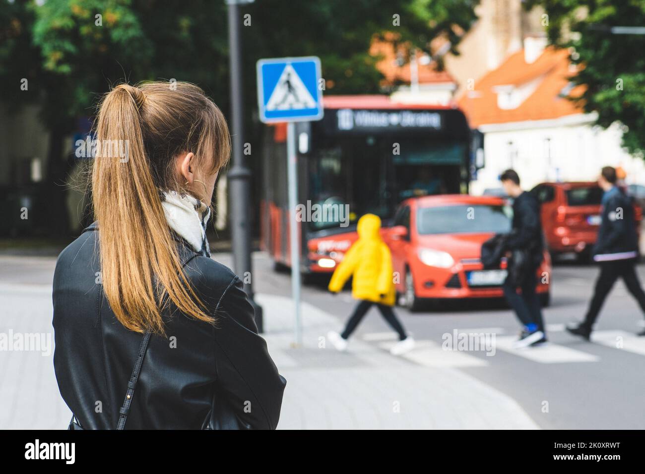 Blonde hair girl waiting at the bus stop on the sidewalk near the road with pedestrian crossing and bus coming in Vilnius, Lithuania Stock Photo