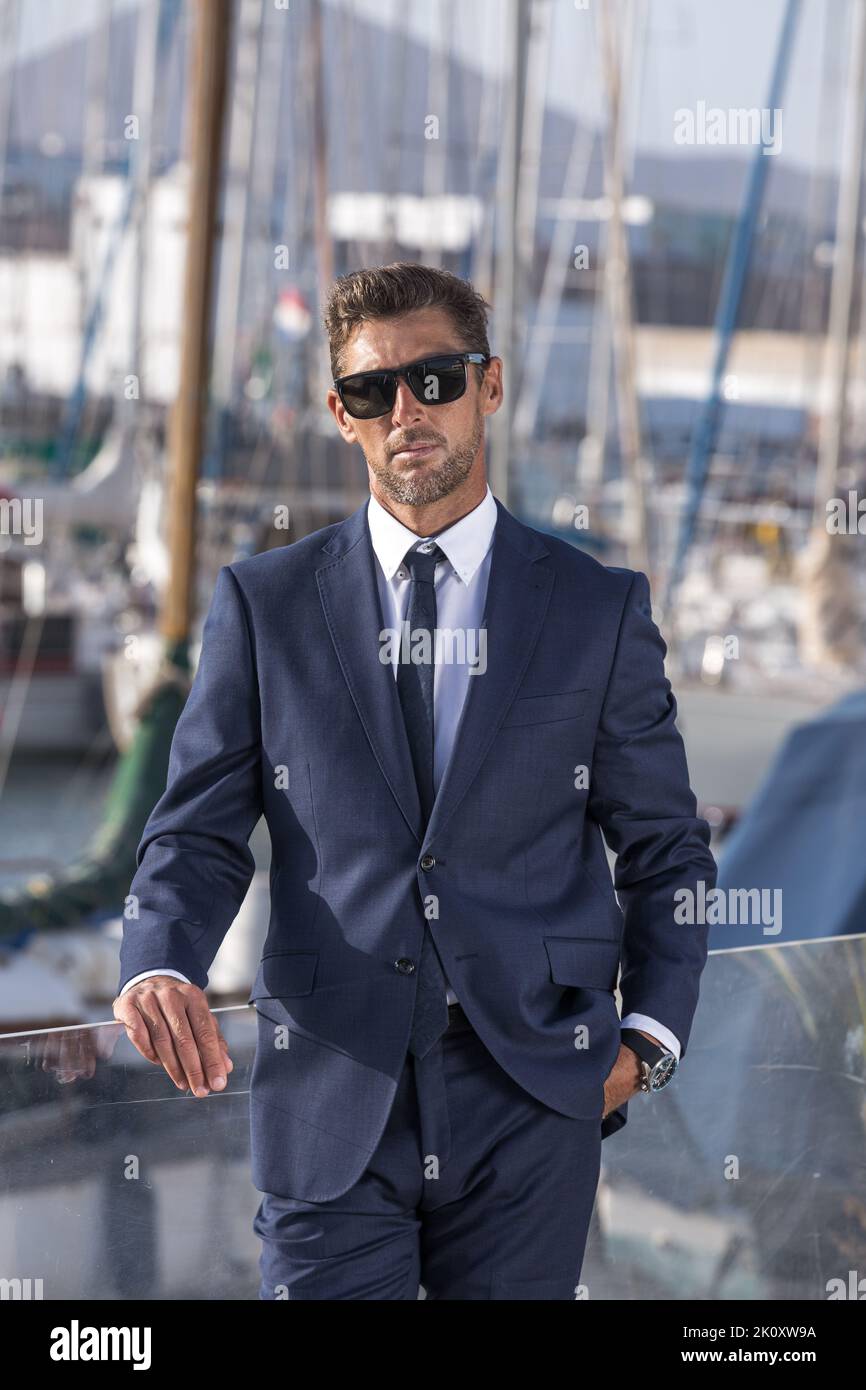 Self assured businessman in suit and sunglasses holding hand in pocket and looking at camera while leaning on fence on blurred background of yachts in Stock Photo