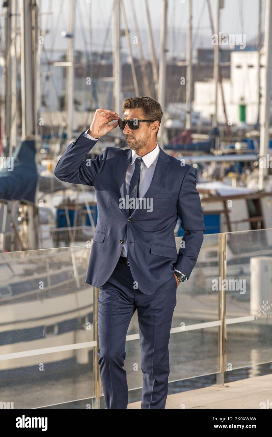 Male entrepreneur with hand in pocket of suit adjusting sunglasses and looking away while standing on embankment against yachts on sunny day in city Stock Photo