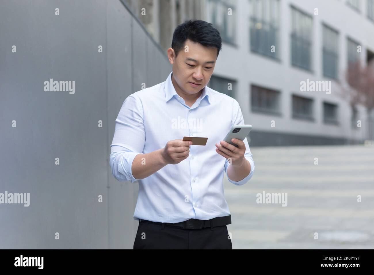 Portrait of a young asian man standing on the street near a modern office center, holding a phone and a credit card in his hands, entering the card number, making online purchases, placing an order. Stock Photo