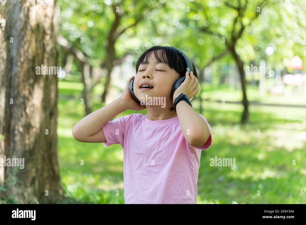 Portrait of child girl listens to music with modern headphones in park outdoors. Happy child enjoying rhythms in listening to music with headphones wi Stock Photo