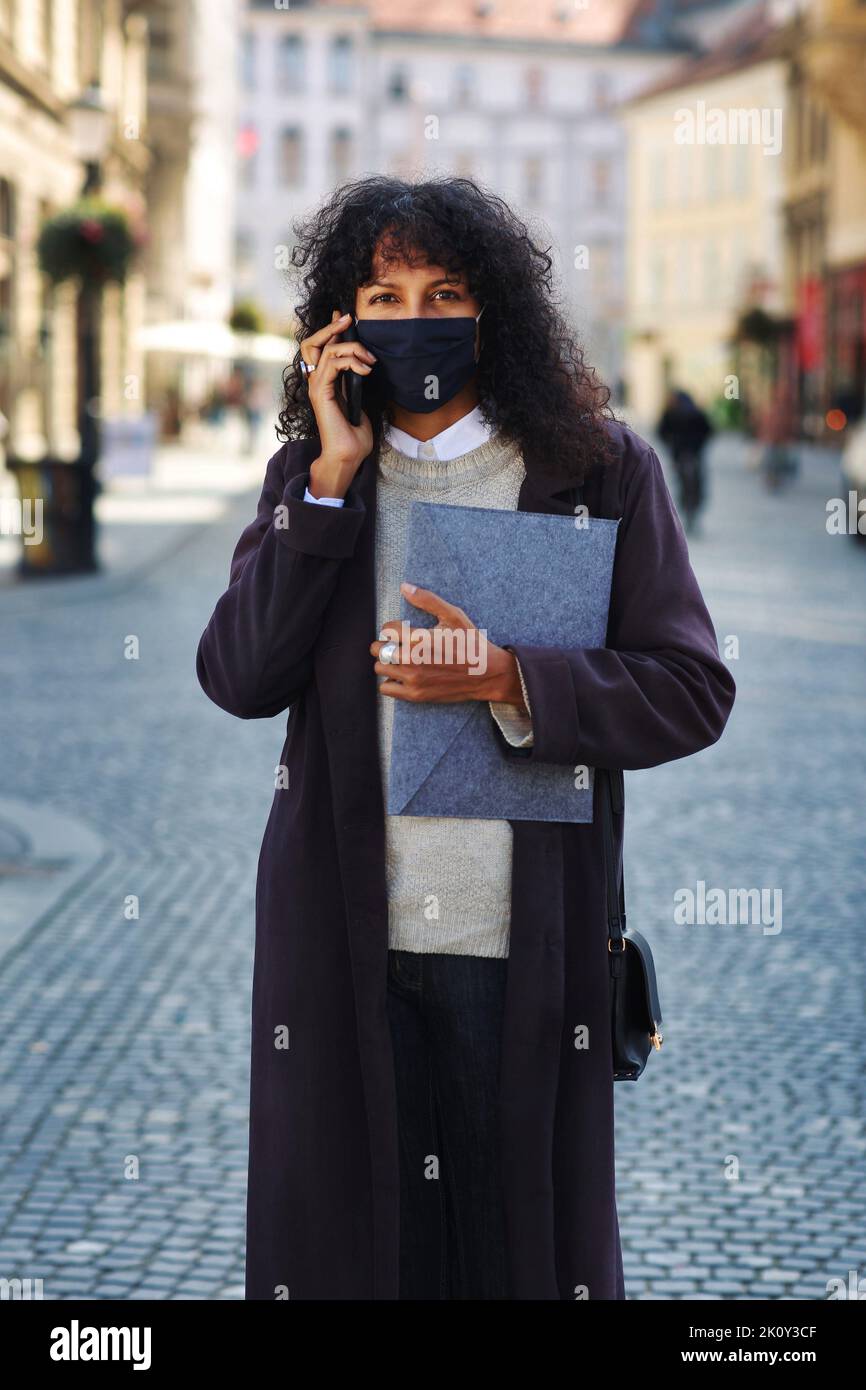 businesswoman outdoors with face mask talking on phone holding folder with documents Stock Photo