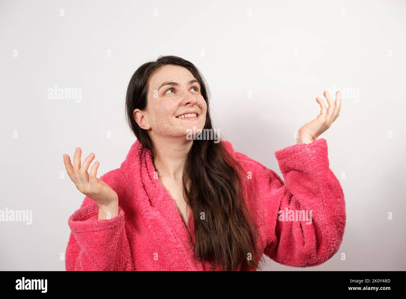 A young brunette woman in a pink bathrobe makes cleansing facial treatments - a mask or scrub to cleanse and nourish the skin. Stock Photo