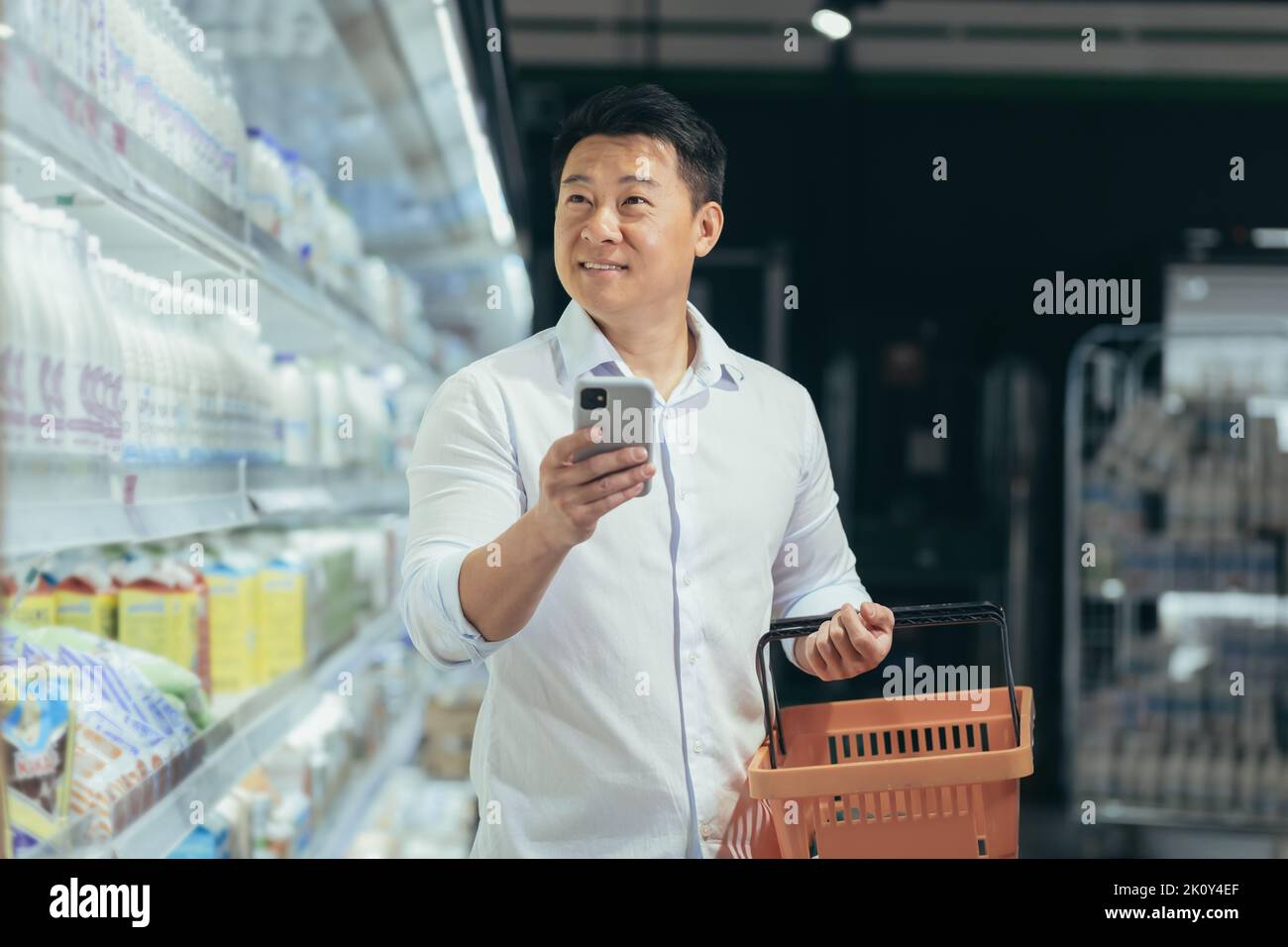 asian man shopper walks around the supermarket with a shopping cart looking at smart phone browsing smartphone in hands search goods or products on the list customer purchaser male in a grocery store. Stock Photo