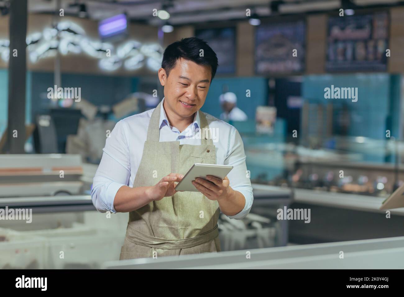 Asian grocery store manager salesman in apron using digital tablet counting goods in supermarket worker employee doing inspection of products regular inventory to control goods online order Stock Photo