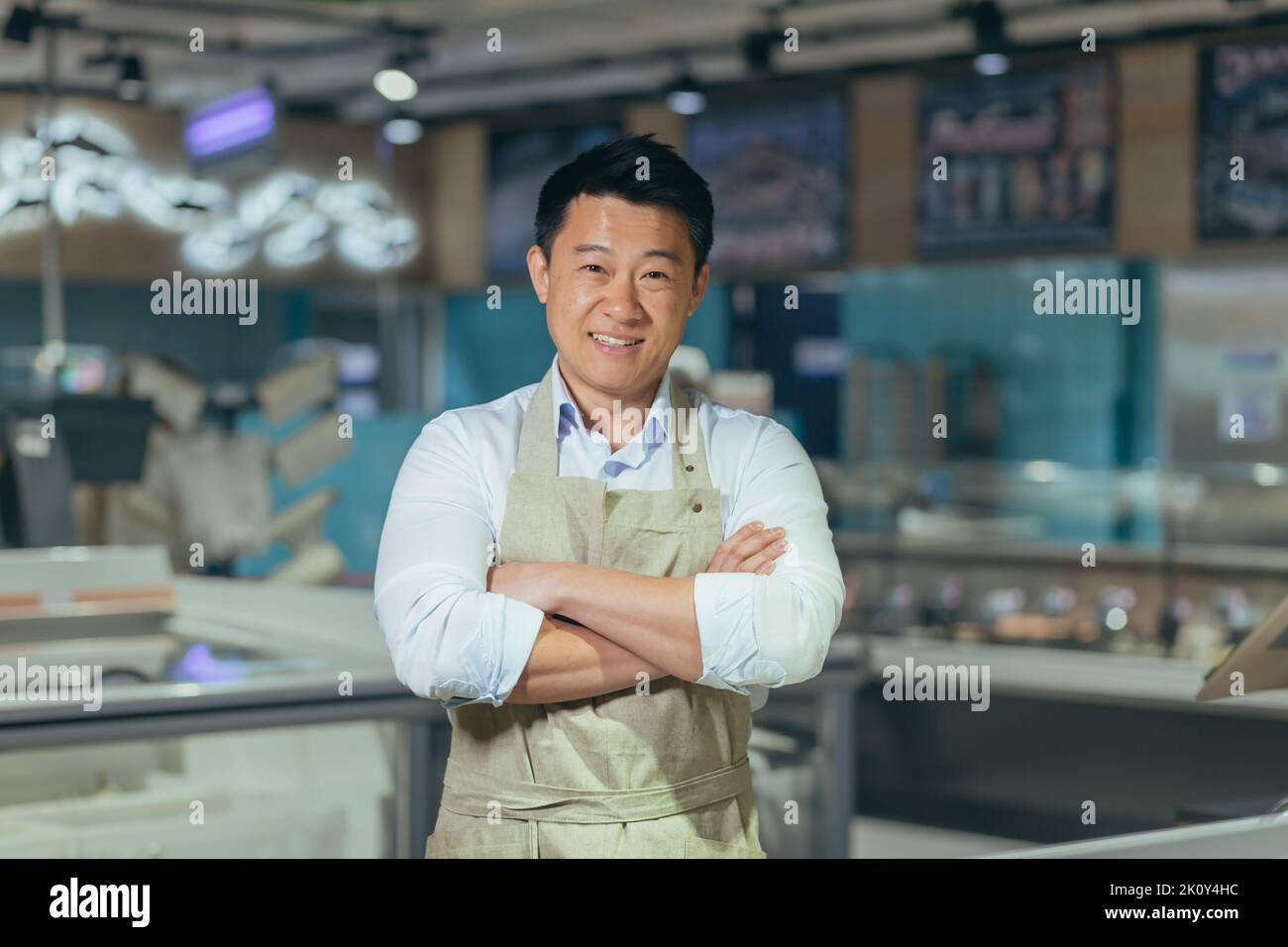 portrait of handsome asian man salesman in apron standing and looking at camera in grocery store supermarket. Male worker seller or small business owner. indoor. food, groceries market Cheerful smile Stock Photo