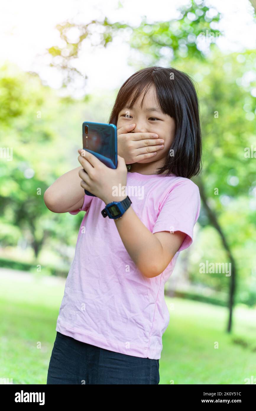 Beautiful asian child girl using mobile smartphone. Little girl is playing in digital game in the green park. Stock Photo