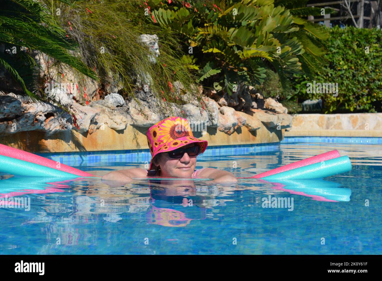 Woman wearing sun hat and shades floating on swim noodles in swimming pool, keeping cool in summer Stock Photo