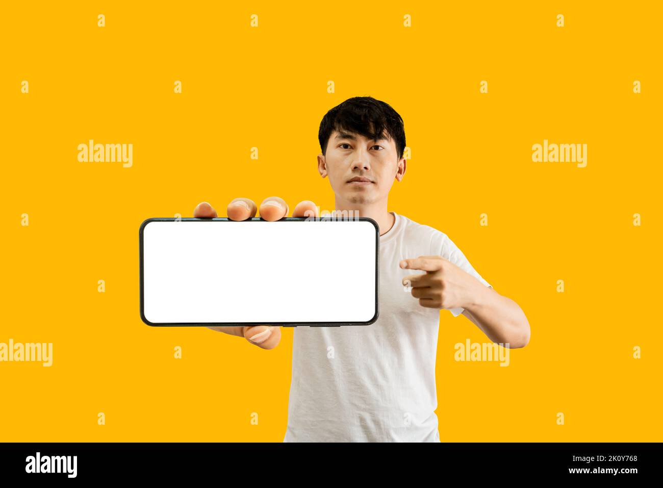 Asian Man Holding Smartphone With White Empty Screen On Yellow Background. Cellphone display Mockup for Mobile App Advertisement. Stock Photo