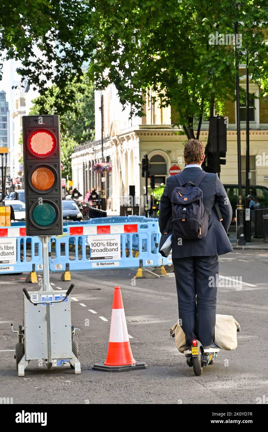 London, United Kingdom - June 2022: Commuter riding an electric scooter with a work bag on the front riding through a red traffic light Stock Photo