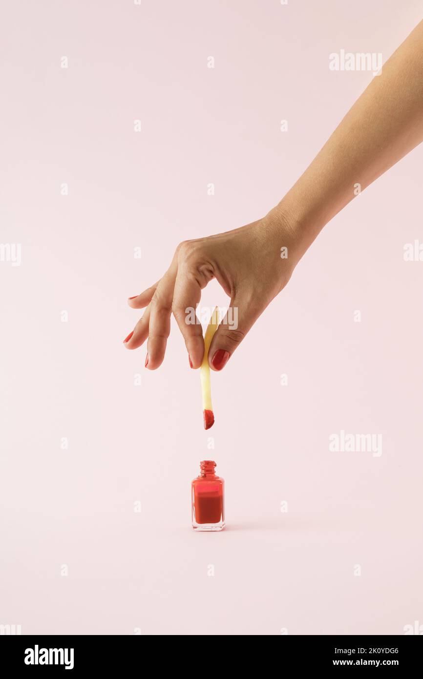 Hand dipping french fried potatoe in red nail polish on pink background. Minimal concept. Stock Photo