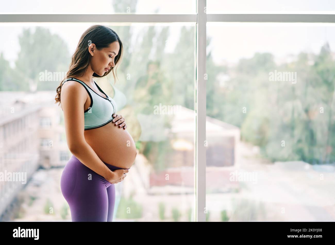 Portrait of young pregnant woman in sportswear standing near window holding hands on her belly with copy space. Pregnancy, expectation and tenderness Stock Photo
