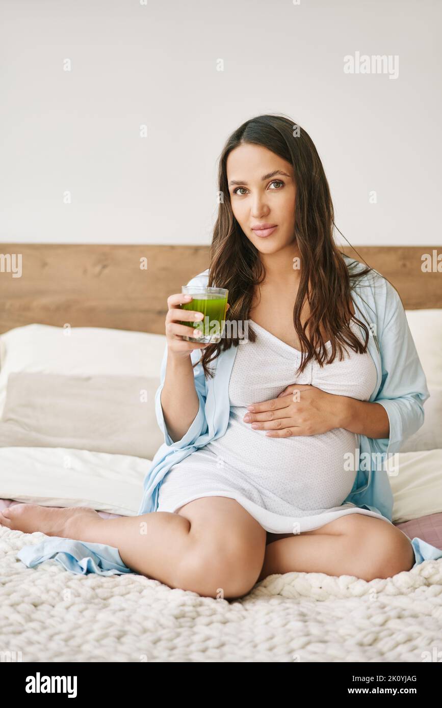 Portrait of young pregnant woman in pajama drinking green smoothie detox juice sitting in bed. Conscious motherhood. Healthy lifestyle concept Stock Photo